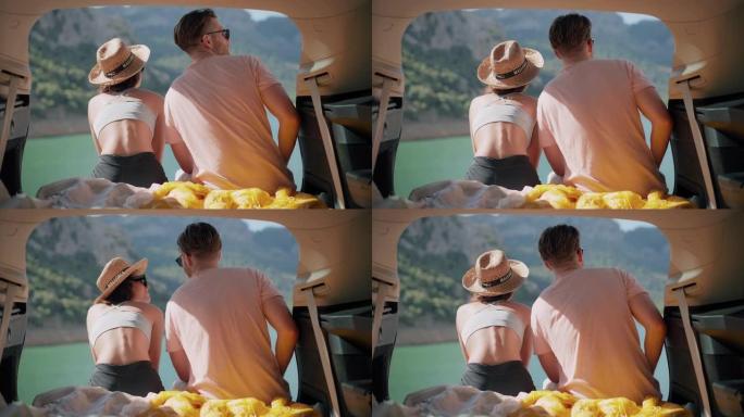 可爱的年轻夫妇坐在美丽的绿松石湖边的汽车后备箱里牵手