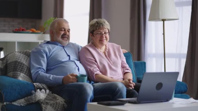 成熟的男人和女人坐在沙发上，通过笔记本电脑与家人进行视频通话，周末在家休息