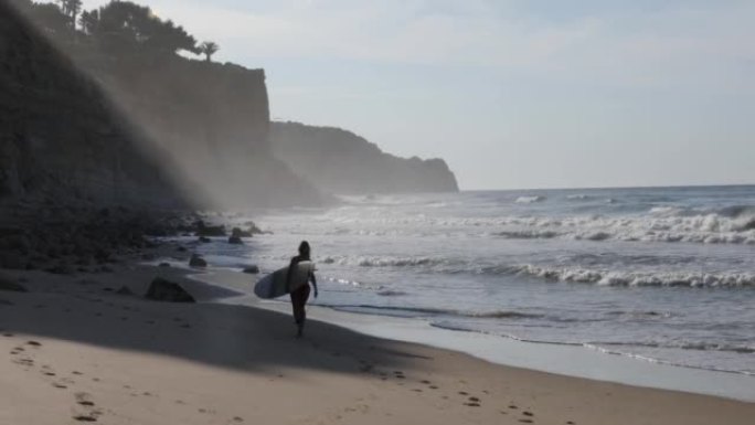 日出时分，年轻女子背着冲浪板沿着空旷的海滩前行