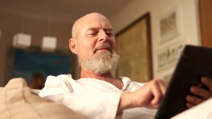 一个自信而快乐的老人在舒适的起居室里咨询他在平板电脑上的投资。