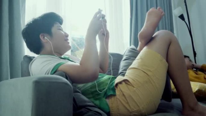 亚洲男孩躺在沙发上，在家窗口附近玩手机游戏，生活方式概念。