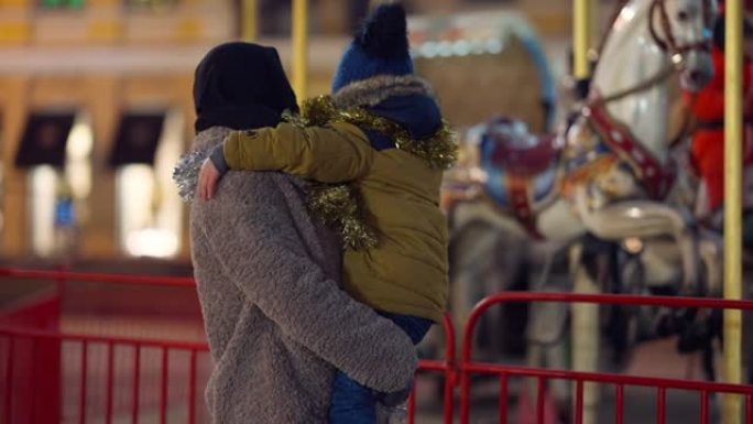 晚上，年轻的中东妇女手中的小男孩在户外欣赏圣诞节旋转木马。快乐过度活跃的儿子和戴着头巾的母亲一起享受
