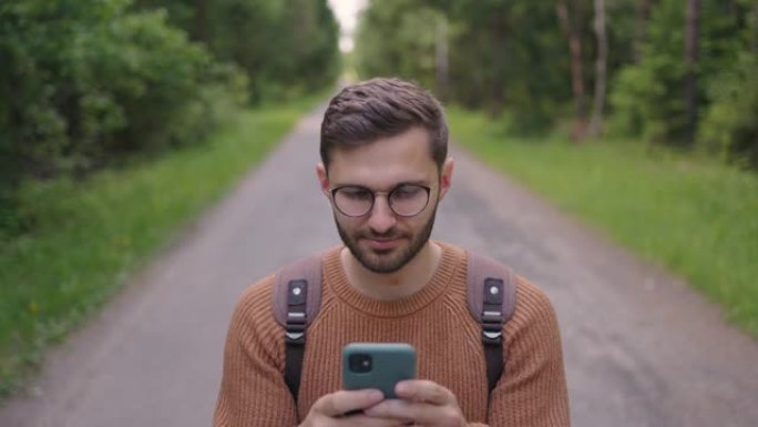 带着胡须的微笑的眼镜男子背着背包在树林中漫步，并以慢动作在手机上打印了一条消息。