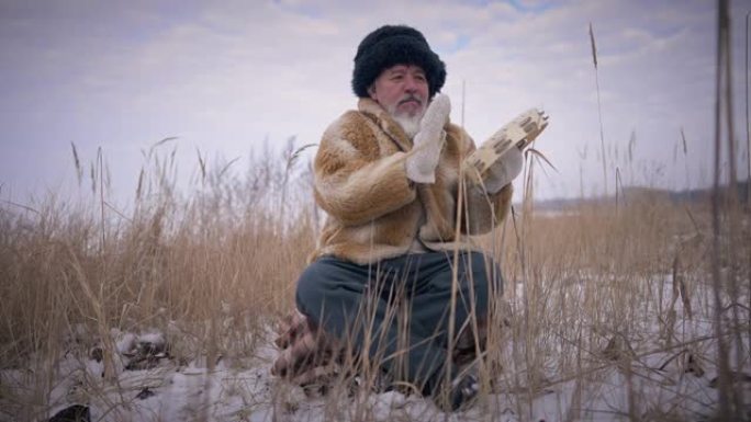 前视图蒙古萨满坐在户外白雪皑皑的冬季草地上慢动作打手鼓。宽阔的肖像自信的亚洲土著男子在美丽的风景。