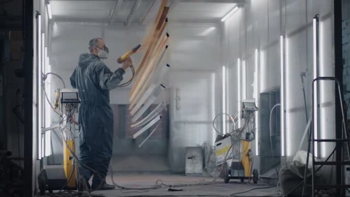 一名身穿防护服和口罩的男子通过喷枪以慢动作将油漆喷洒在钢制零件上。