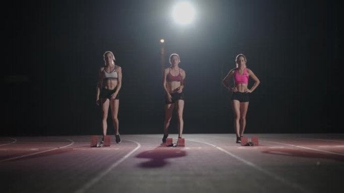 前视图三名女跑步者准备在体育场以慢动作在黑暗的灯光下奔跑