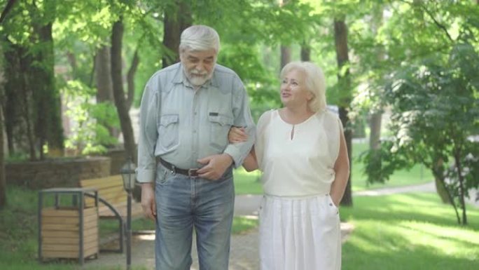 无忧无虑的高级丈夫和妻子在阳光明媚的公园漫步和交谈的肖像。快乐的老高加索男人和女人手牵着手走在户外。