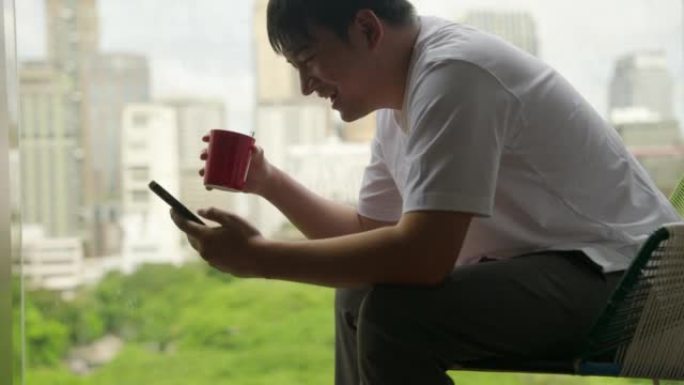 亚洲人坐在一栋享有城市美景的建筑的阳台上喝咖啡。