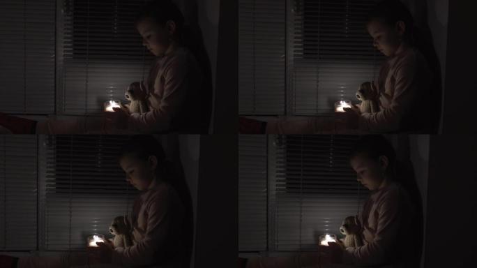 窗边黑暗房间里的一个女孩手里拿着蜡烛和泰迪熊