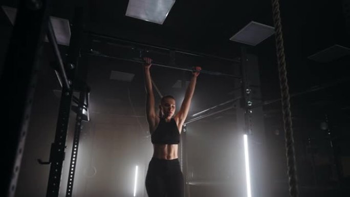 运动女子在健身房的横杆上做引体向上，晚上在黑暗的健身馆里独自训练