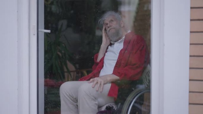 坐在轮椅上的体贴悲伤的残疾老人坐在玻璃门后面，在雨天望着外面。家里心烦意乱的高加索残疾人退休人员的肖