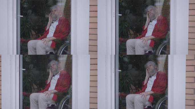 坐在轮椅上的体贴悲伤的残疾老人坐在玻璃门后面，在雨天望着外面。家里心烦意乱的高加索残疾人退休人员的肖