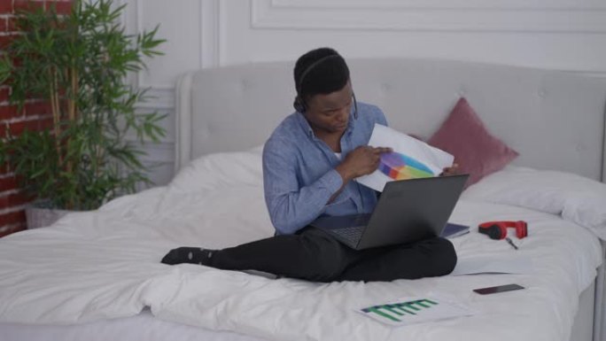 坐在床上的耳机里自信的非洲裔美国年轻人的宽幅肖像，指着笔记本电脑上的视频聊天。家庭办公室内远程工作便