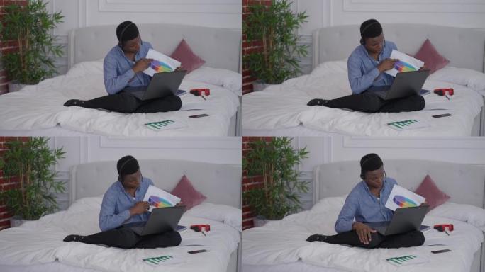 坐在床上的耳机里自信的非洲裔美国年轻人的宽幅肖像，指着笔记本电脑上的视频聊天。家庭办公室内远程工作便
