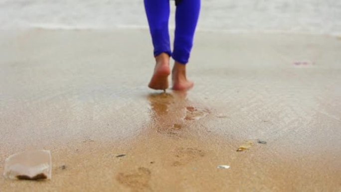 一个年轻女孩光着脚走路，留下脚印的特写镜头，抚摸着沙滩上溅起的水，海浪向她走来。