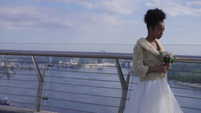 穿着婚纱的沮丧美丽的非洲裔美国妇女把新娘花束放在桥扶手上，慢动作离开。悲伤的新娘在婚礼那天走开了。失