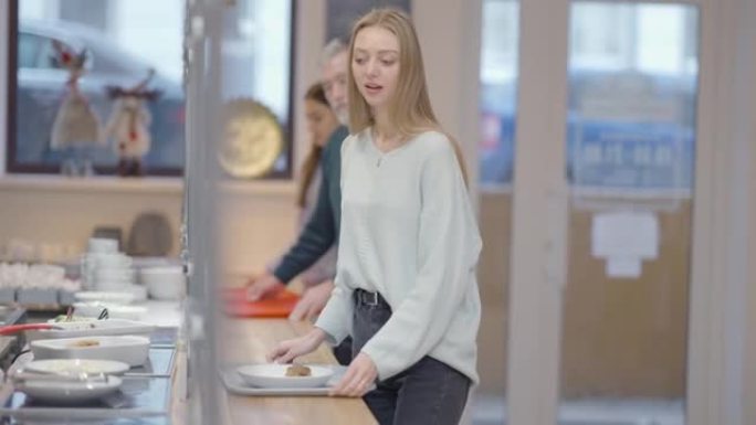 积极苗条的年轻女子在自助餐厅点菜，慢动作排队。微笑着美丽的白人女士在午餐时用餐。背景下模糊的人。