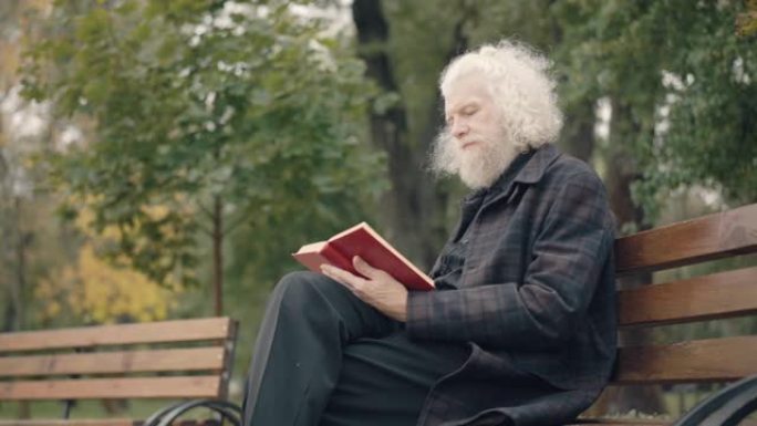 秋天公园读书的集中老人坐在长凳上的侧视图。吸收了白人男性退休人员，退休后在户外享受业余爱好。生活方式