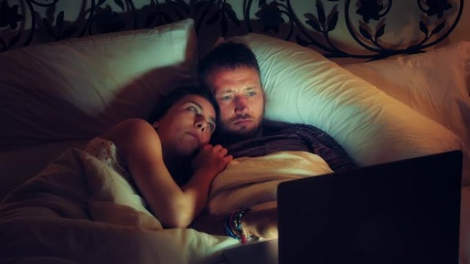年轻夫妇在床上看电影。使用笔记本电脑，玩得开心