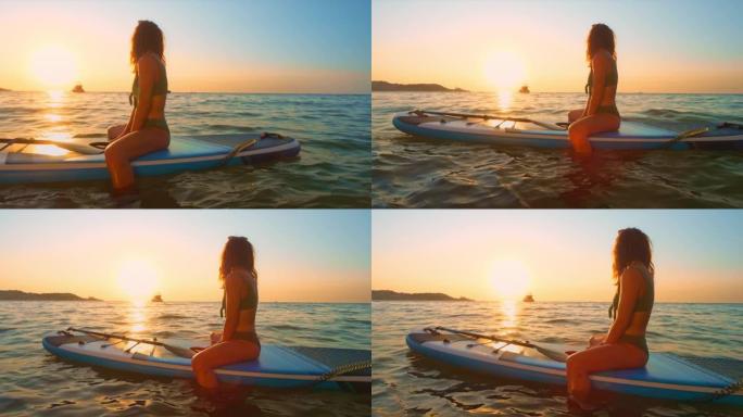 SLO MO女人坐在桨板上，看着海面上的日落
