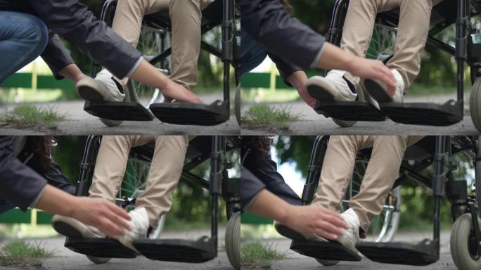 无法辨认的女人在慢动作中调整轮椅上残疾儿童的腿。年轻的高加索母亲在夏季公园户外照顾截瘫的儿子。