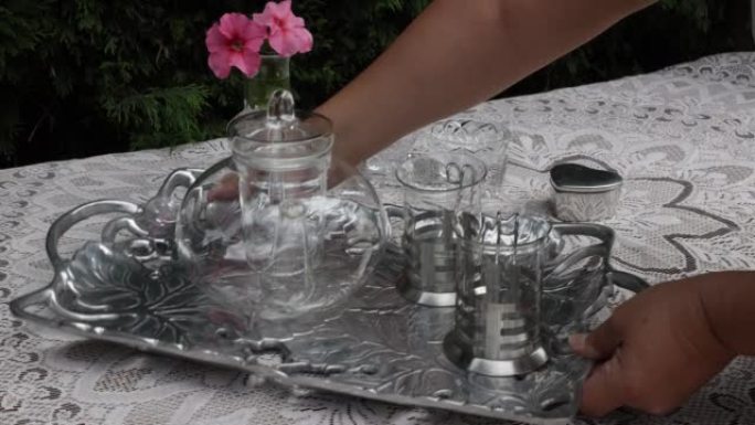 成年女性手将一个金属托盘和一个玻璃茶壶和两个杯子放在桌子上