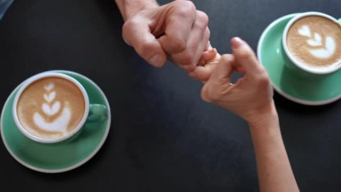 顶视图咖啡桌，男女双手示意小指发誓。特写无法辨认的高加索爱好男女手牵手坐在室内咖啡馆约会。爱与团结。