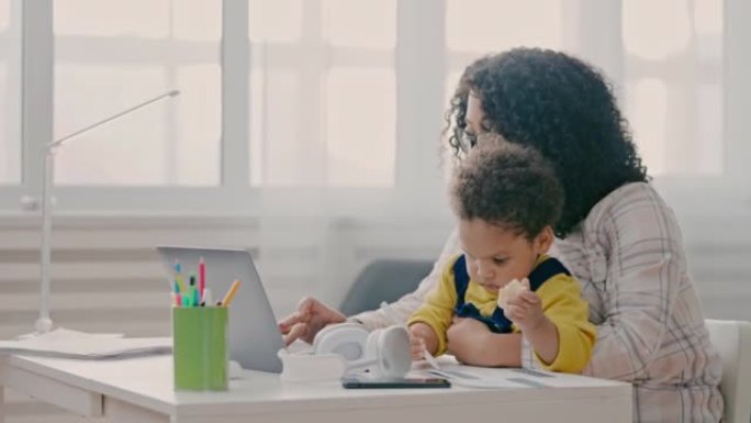 忙碌的妈妈抱着蹒跚学步的孩子，在笔记本电脑上工作，育儿挑战，照顾孩子