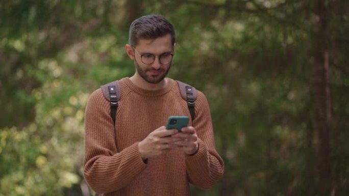 一个背着背包和手机的男性旅行者正穿过森林，试图捕捉信号。在森林地区的手机上搜索信号。