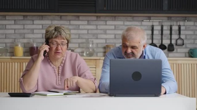 退休妇女正在打电话给公用事业公司并咨询付款，她的丈夫坐在附近，用笔记本电脑浏览互联网