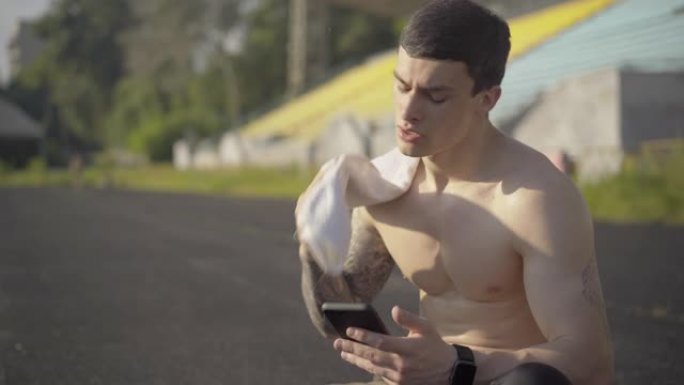 出汗运动员的中间镜头用智能手机用白毛巾擦脸。健康英俊的白人男子的肖像在运动场上晨练后休息。
