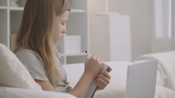 十几岁的女孩正在家里用笔记本电脑听老师的在线讲座，在字帖中写作，在流行病期间为学生提供电子学习