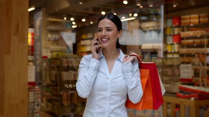 美丽的年轻女子带着纸袋离开商店，一边兴高采烈地在智能手机上聊天