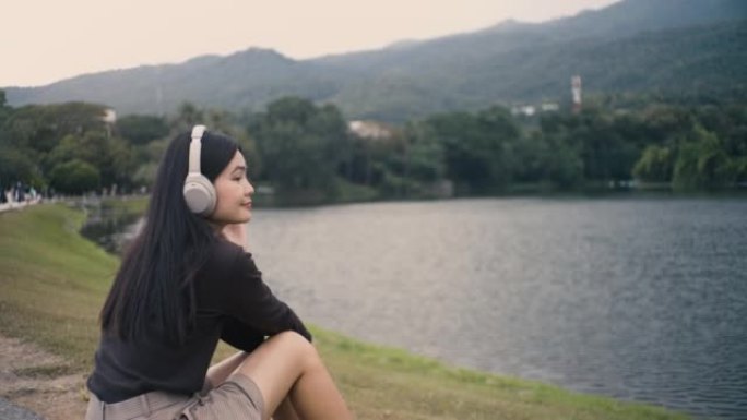 亚洲女孩戴着耳机听音乐，脸上挂着快乐的微笑，坐在公园的座位上，有湖泊和山脉。