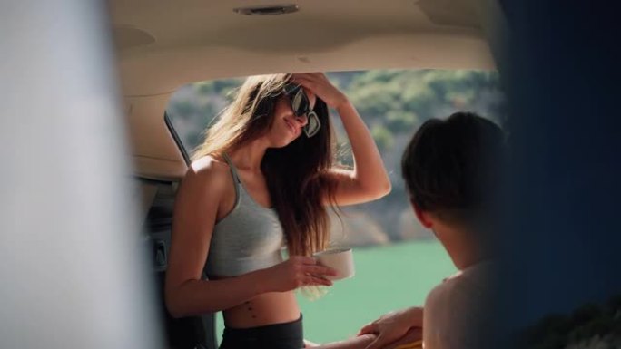 可爱的年轻夫妇坐在美丽的绿松石湖边的汽车后备箱里喝茶
