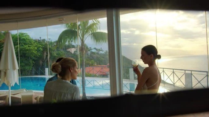 女性朋友在度假别墅聊天喝酒。热带度假。透过阴影百叶窗