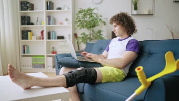 年轻的卷发男子在笔记本电脑上工作，正在接受康复治疗，膝盖受伤
