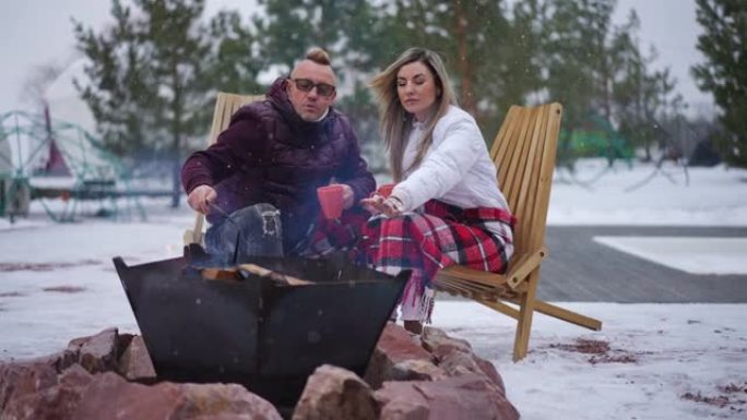 实时摄像机放大，让自信放松的白人夫妇坐在篝火旁聊天移动的柴火。白雪皑皑的冬日快乐无忧无虑的夫妻享受生