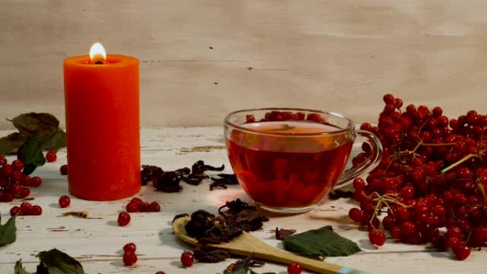 在白色的木桌上放红茶和燃烧的蜡烛的杯子。木制乡村木板上的红色荚果浆果中的天然茶。自然疗法愈合输液。抗