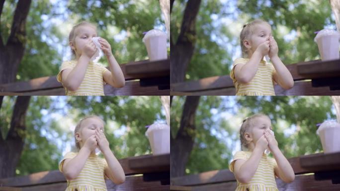 小女孩吃完后用餐巾纸擦脸，桌上放着奶昔。可爱的小女孩坐在公园长椅上，用纸巾擦脸的特写镜头。