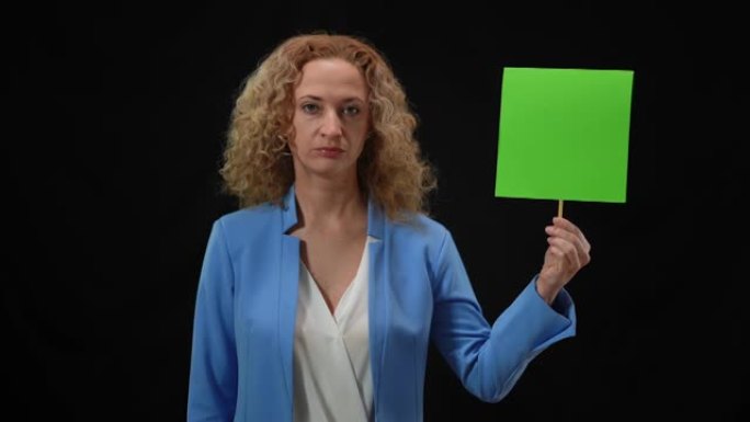肖像女子拿着绿色标志摇头，表情严肃。优雅自信的白人公众人物在黑人背景下拒绝否认。