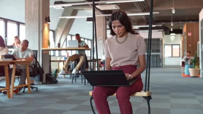 DS年轻女子坐在办公办公室的秋千上，在笔记本电脑上工作