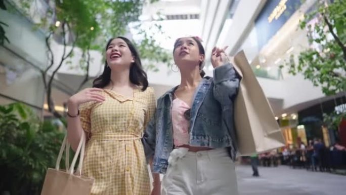两个年轻的购物狂亚洲女性在购物大道中间散步时展示了美好的时光，手里拿着很多纸购物袋，女性社会，外出女