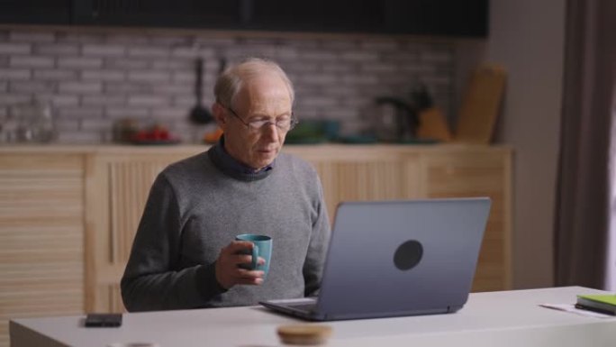 爷爷在笔记本电脑上通过视频通话聊天，独自坐在家庭厨房里，在大流行期间进行远距离交流