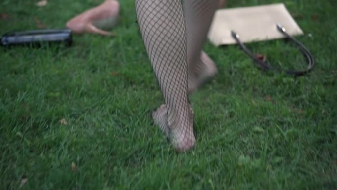 跟踪镜头赤脚苗条的年轻醉酒女子在渔网长袜上行走在绿色的草地上通过空的酒精瓶散落的高跟鞋和钱包。醉酒的