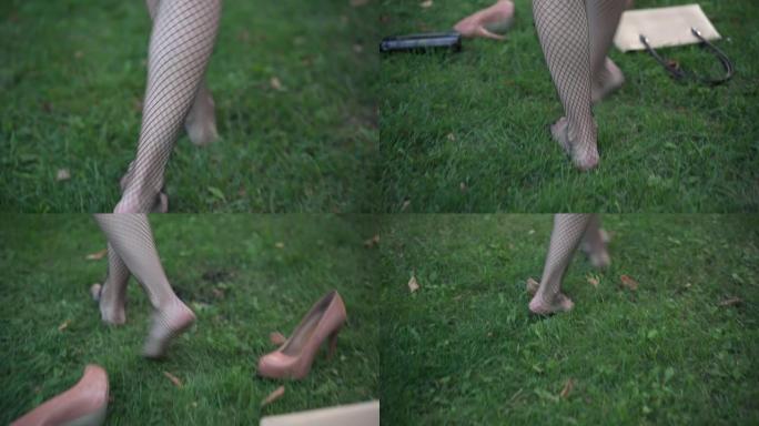 跟踪镜头赤脚苗条的年轻醉酒女子在渔网长袜上行走在绿色的草地上通过空的酒精瓶散落的高跟鞋和钱包。醉酒的