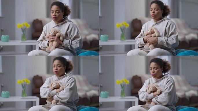 沮丧的年轻非洲裔美国妇女拥抱玩具兔子的肖像坐在室内。表情沮丧的孤独悲伤美丽女士的中景。绝望和孤独。