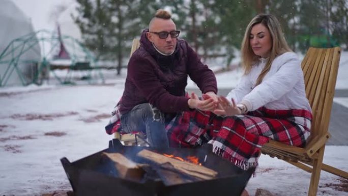 下雪天，放松的成年夫妇坐在户外，在篝火旁聊天。自信的高加索男人和女人享受寒冷的冬日休息聊天的宽幅肖像