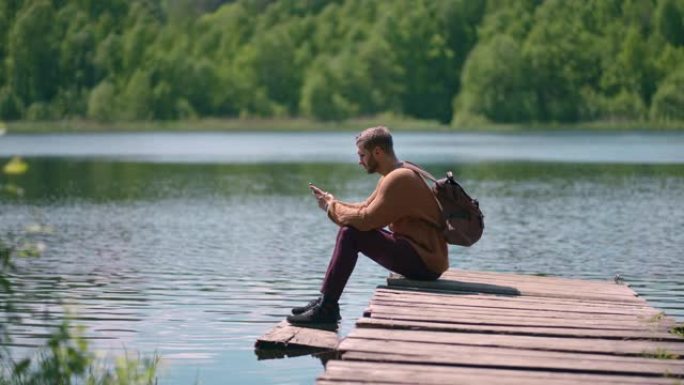 一位戴着眼镜、背着背包、蓄着胡须的年轻男子坐在湖边的一个木制码头上，用慢镜头打印出一条信息，用手机铺