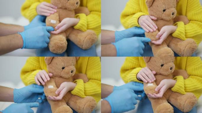儿科医生用儿童手在泰迪熊上应用医疗贴片的特写。无法辨认的医生在室内儿科诊所安抚病人。医院护理和医学概